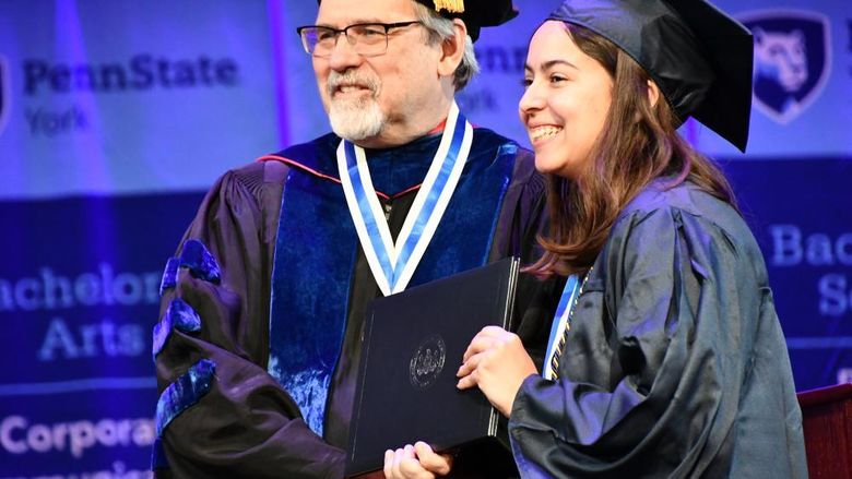 Man shakes hands with young lady accepting diploma, dressed in regalia. 