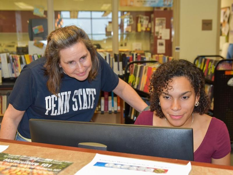 Woman wearing a Penn State T-shirt looking over the shoulder of a Latino female student working at a computer.
