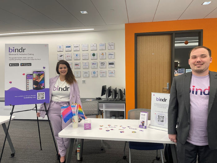 Two people stand in front of display boards at tables promoting an app called Bindr