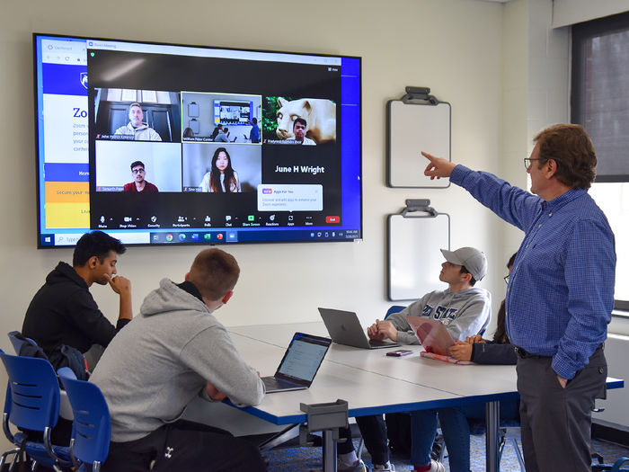 Faculty member pointing at screen with students seated at a table with laptops and others pictured on screen remotely