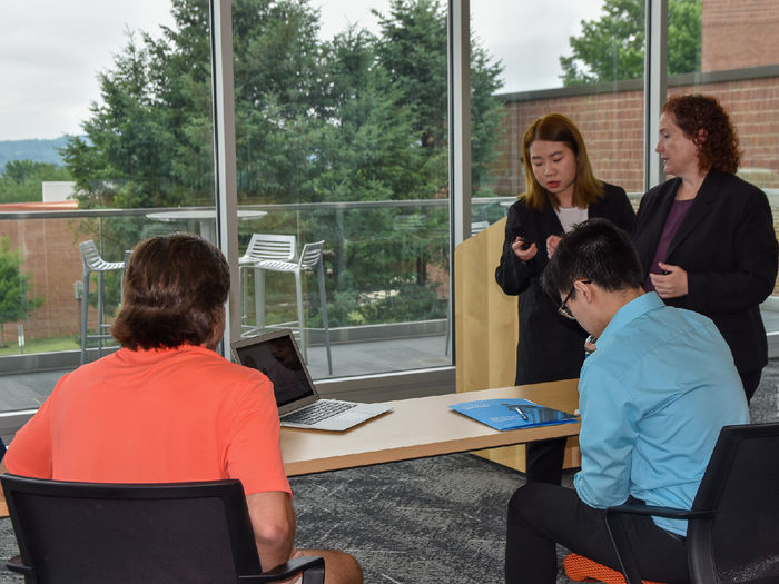 Young women and female faculty member discuss a presentation in front of two male students seated at a table 