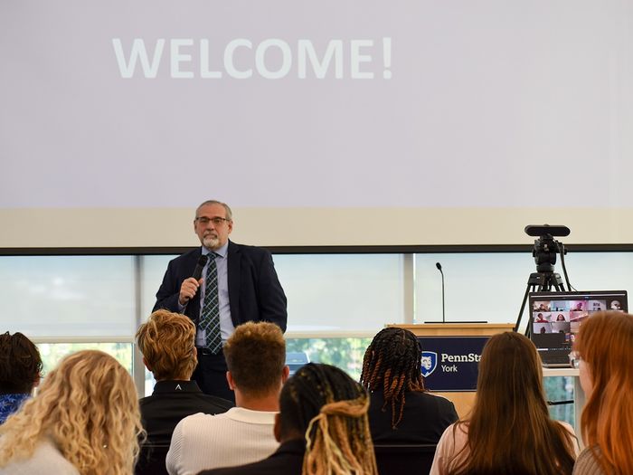 Man speaking into a microphone in front of a group of college students. The word "welcome" is on the screen behind him.