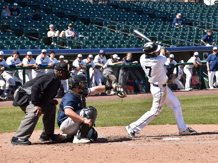 Umpire and catcher positioned behind a student in blue and white baseball uniform swinging a bat