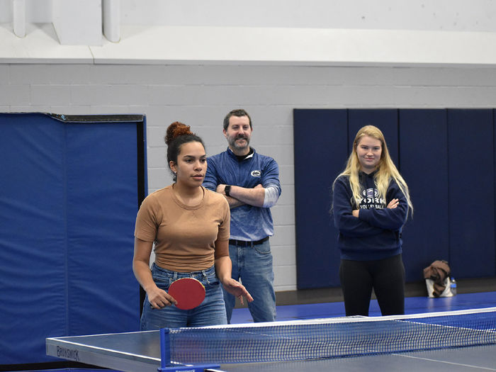 A Latino female college student plays ping pong while an older male and female college student watch. 