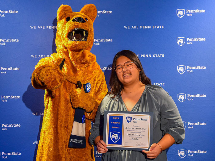 Female make student wearing glasses and holding an award along with the Nittany Lion character