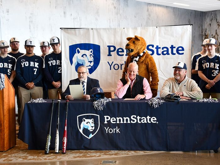 A baseball team stands around three men sitting at a table, with the Nittany Lion mascot behind them.