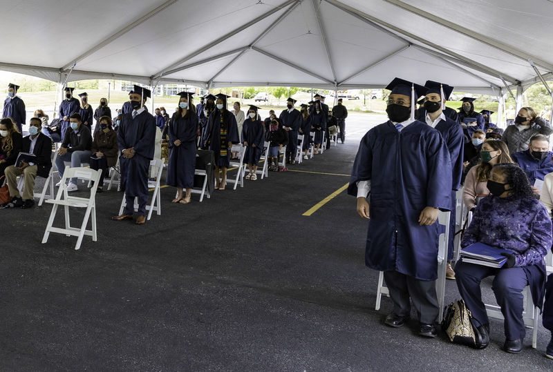 Students dressed in caps and gowns and wearing masks stand while audience members remain seated.