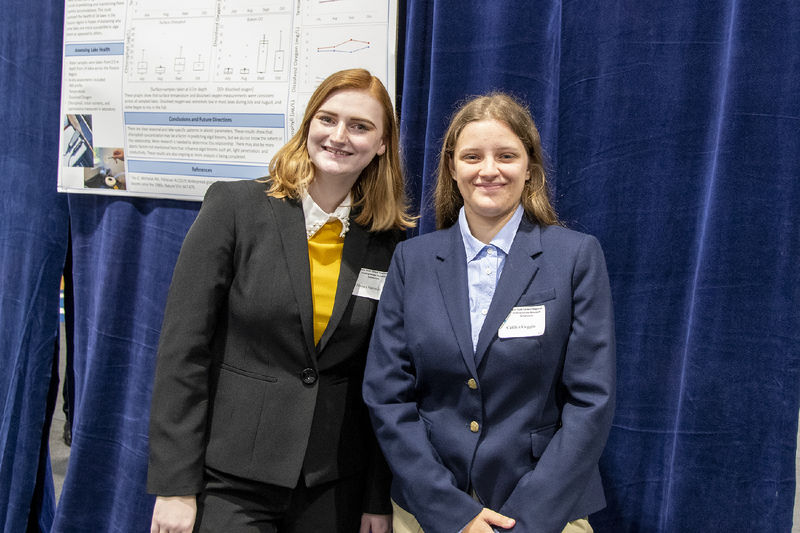 Two students wearing suits pose for a photo in front of a research poster.