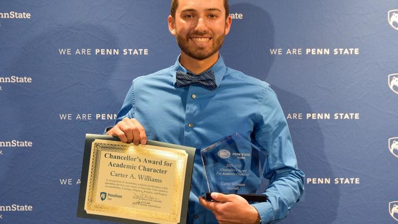 Caucausian male with dark hair holding award certificate while standing in front of a blue Penn State backdrop.