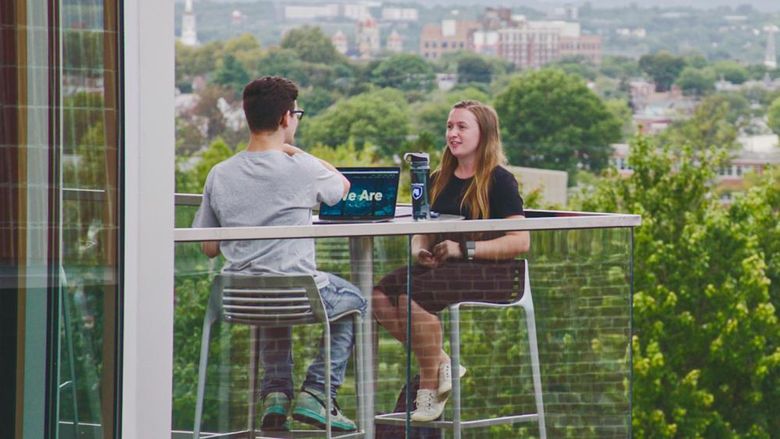 Male and female student sitting on balcony with York City skyline in the background.