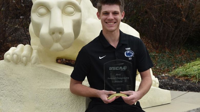 Male student holding golf award as he sits  in front of the Penn State NIttany Lion Shrine 
