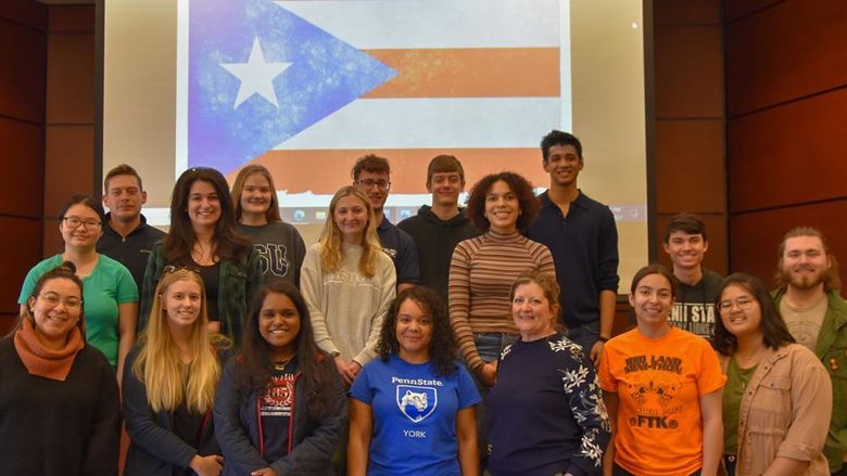Group of 18 students and two staff members on a stage in front of the flag od Puerto Rico