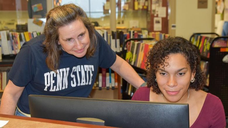 Woman wearing a Penn State T-shirt looking over the shoulder of a Latino female student working at a computer.