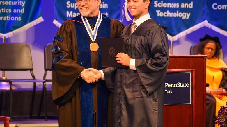 Older man and student in commencement regalia on stage shaking hands