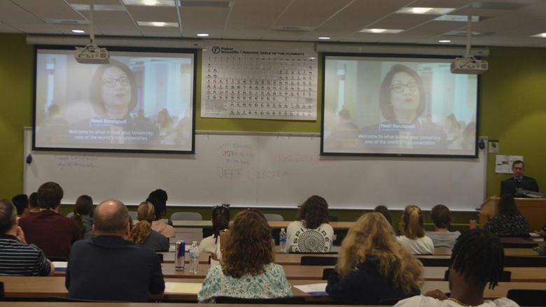 Photo from the back of a room with people seated and watching a screens during a program.