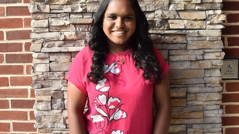 A person wearing a hot pink flowered top, standing against a rock wall.