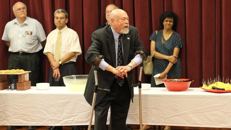 Older man wearing leg braces and speaking, several people in the background