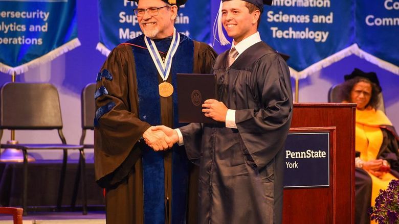 Male student in cap and gown shaking hands with older man wearing glasses and dressed in academic regalia