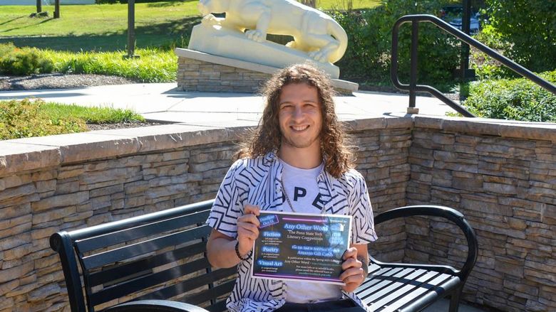 Young man with should length curly hair sitting on a bench holding a sign to promote an event.