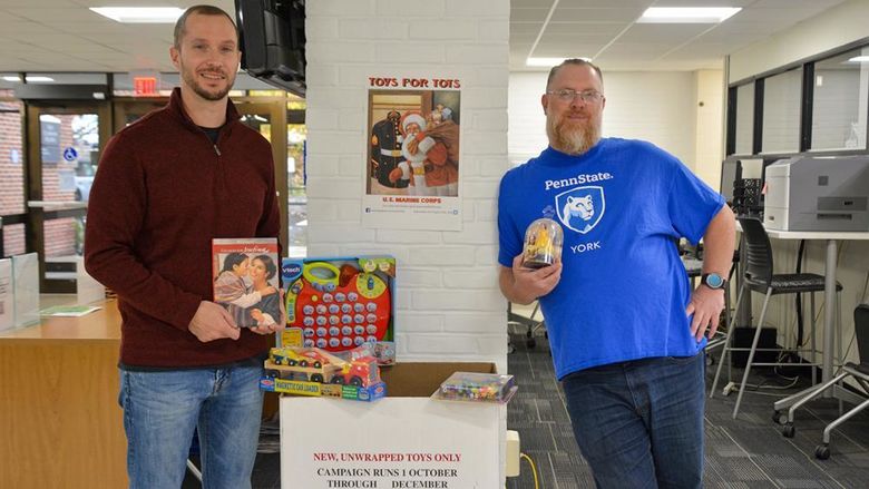 Two male adult students stand holding toys near Toys for Tots collection box