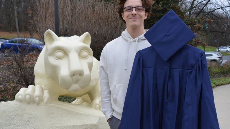 A male student holding academic regalia and standing next to the Nittany Lion Shrine at Penn State York