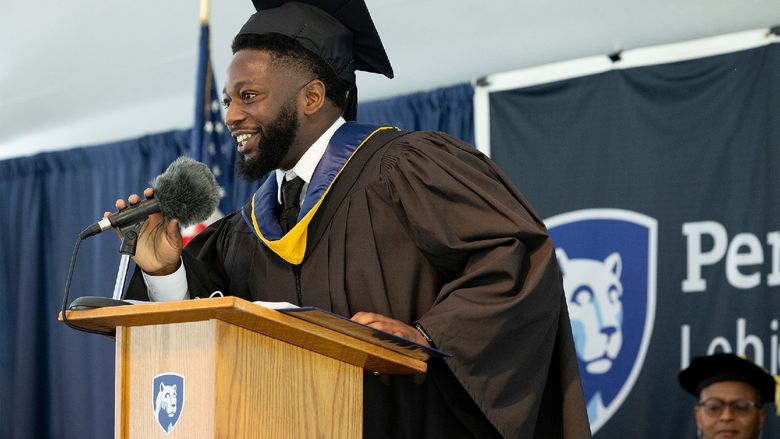 Abu Fofanah wearing commencement regalia standing at a podium speaking.