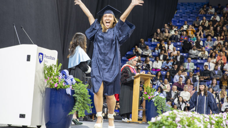 Student wearing cap and gown jumps for joy crossing the stage at commencement