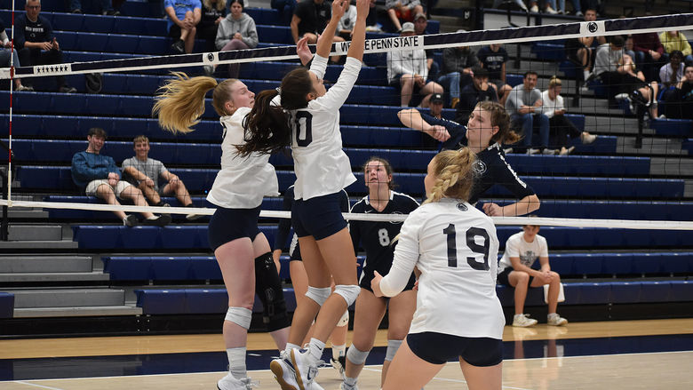Four women's volleyball players at the net with two going up for a hot and the other defending