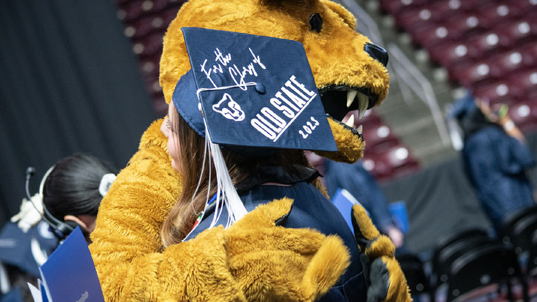 The Nittany Lion hugs a graduate in a cap that reads "For the Glory of Old State 2023"