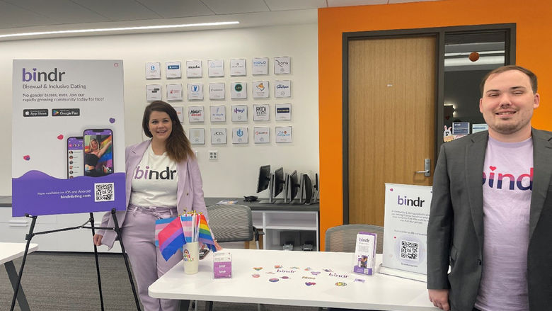 Two people stand in front of display boards at tables promoting an app called Bindr