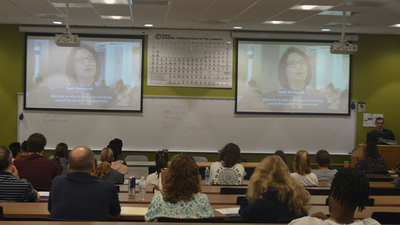 Photo from the back of a room with people seated and watching a screens during a program.
