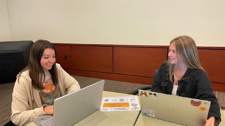 Two female students with long hair working at laptops at a table