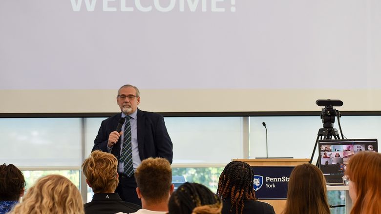 Man speaking into a microphone in front of a group of college students. The word "welcome" is on the screen behind him.