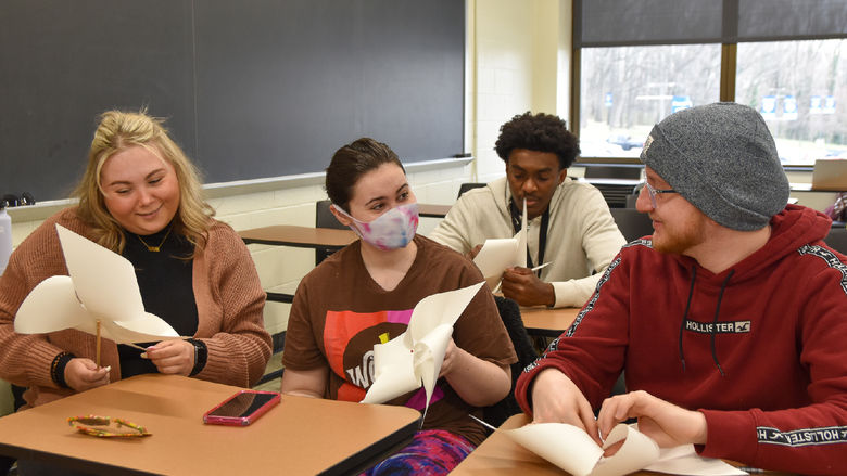 Four students seated at desks, two females and two males, in a classroom making paper pinwheels.