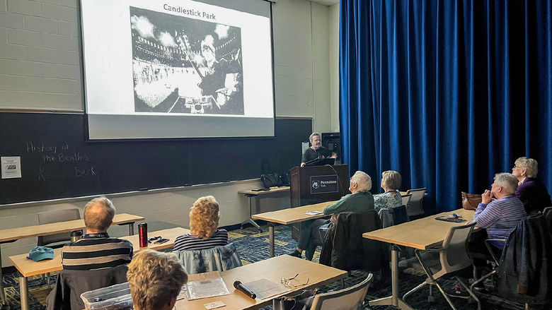 Back view of a classroom of older people at desks and a photo on the screen with instructor at front