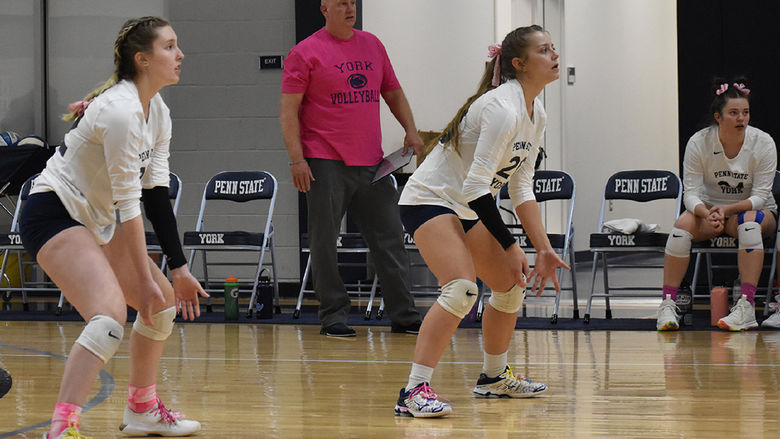 Two women's volleyball players on the court and one on the bench, male coach in the background wearing pink shirt.