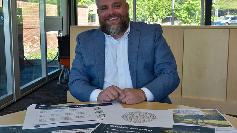 Bearded white male dressed in a suit showing off materials on the desk in front of him.