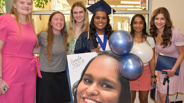 Six female students stading together, with one at the center wearing a graduation cap and gown.