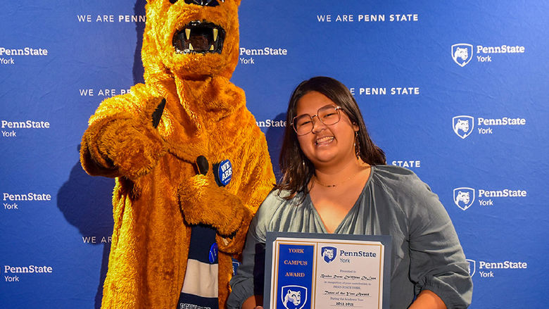 Female make student wearing glasses and holding an award along with the Nittany Lion character