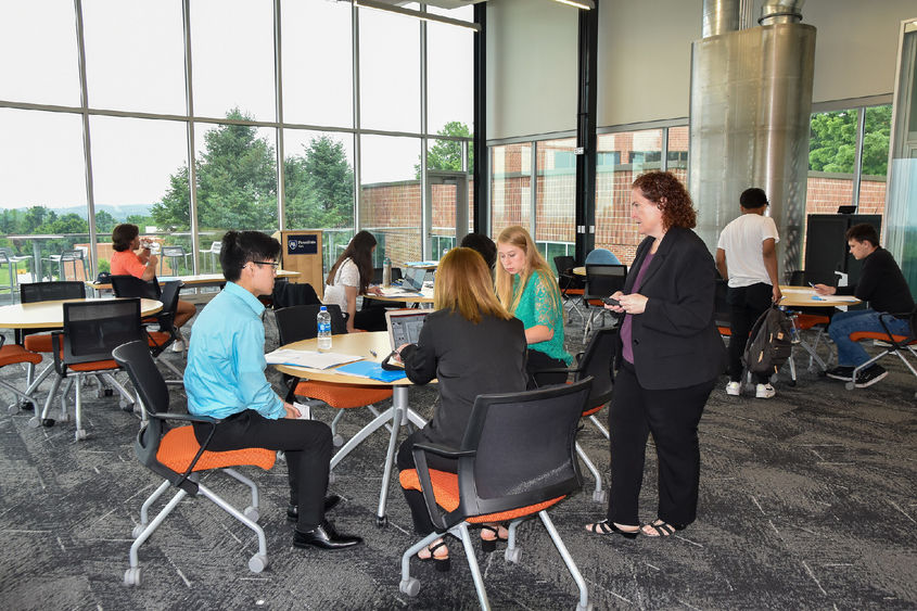 Faculty member  and groups of students arouind tables discussing work