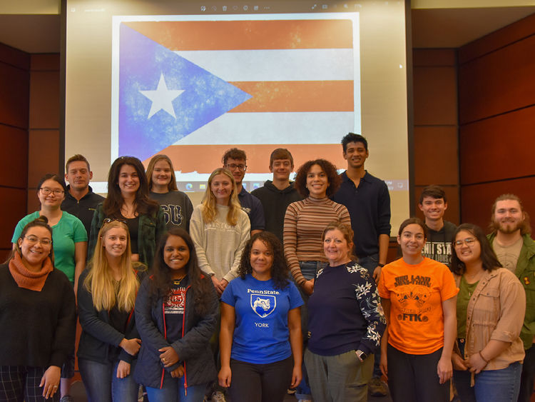Group of 18 students and two staff members on a stage in front of the flag od Puerto Rico