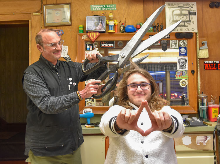 Two males, one older and one young.  Older male is holding large scissors and pretending to cut younger male's hair who is sitting in a barber's chair.