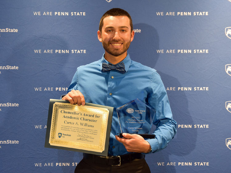 Caucausian male with dark hair holding award certificate while standing in front of a blue Penn State backdrop.