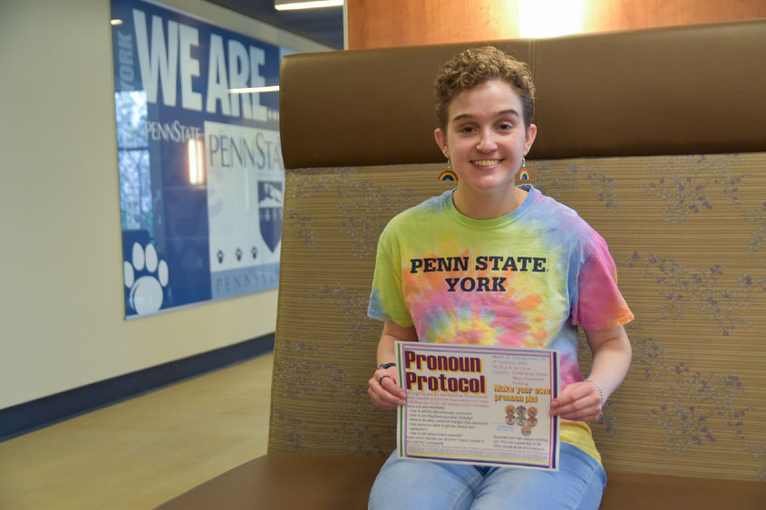 Seated short-haired female wearing colorful shirt hold program sign for Pronoun Protocol program