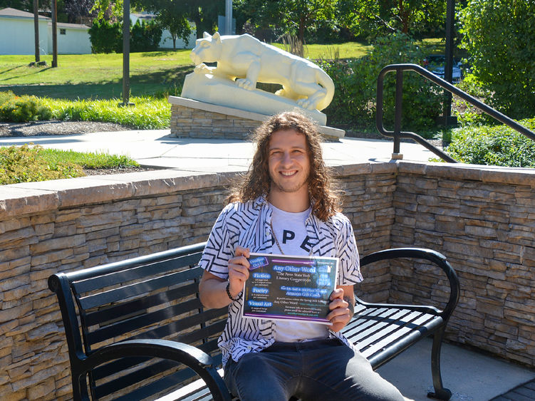 Young man with should length curly hair sitting on a bench holding a sign to promote an event.