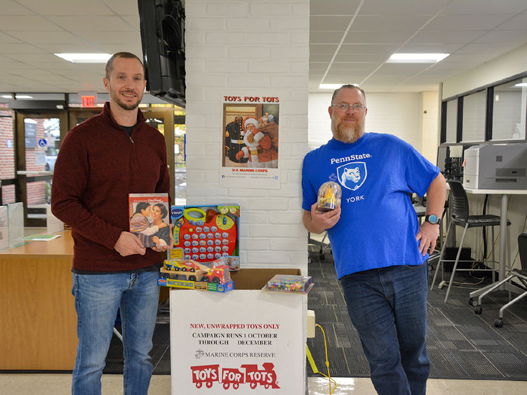 Two male adult students stand holding toys near Toys for Tots collection box