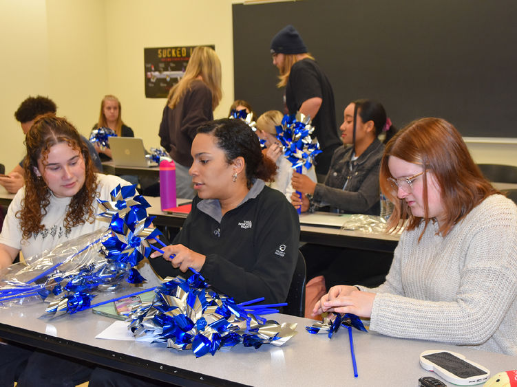 Three females college student at a table assembling pinwheels, male and female classmates in the background doing the same