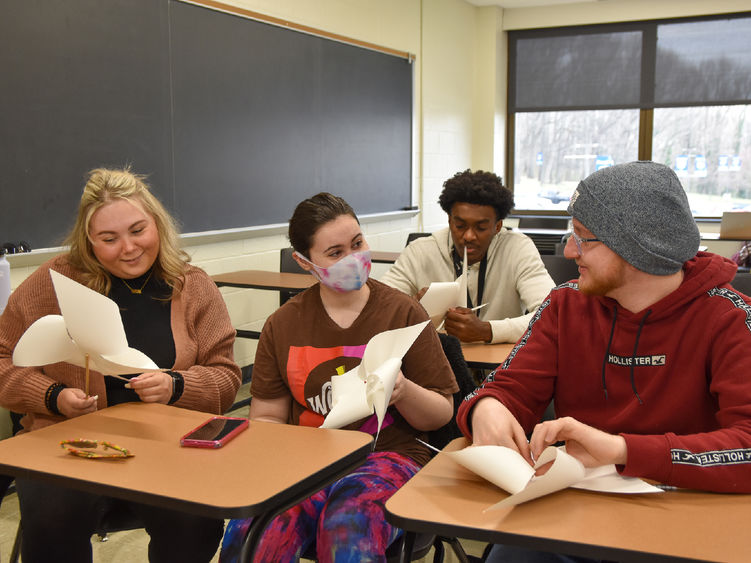 Four students seated at desks, two females and two males, in a classroom making paper pinwheels.