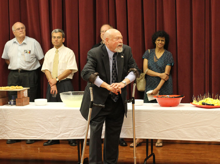 Older man wearing leg braces and speaking, several people in the background