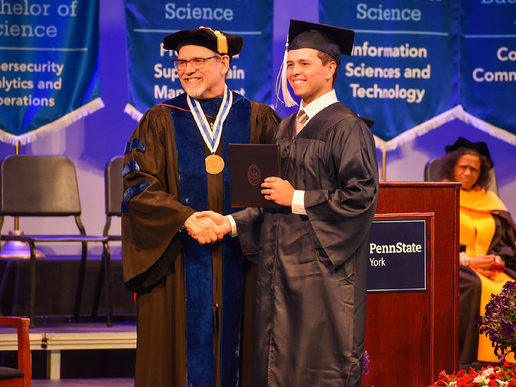 Male student in cap and gown shaking hands with older man wearing glasses and dressed in academic regalia